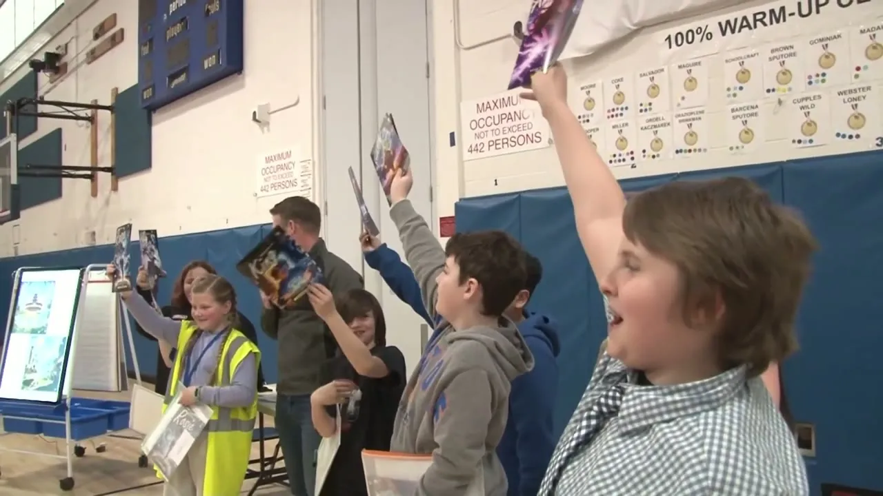Students in a school gym holding their printed comic books above their heads